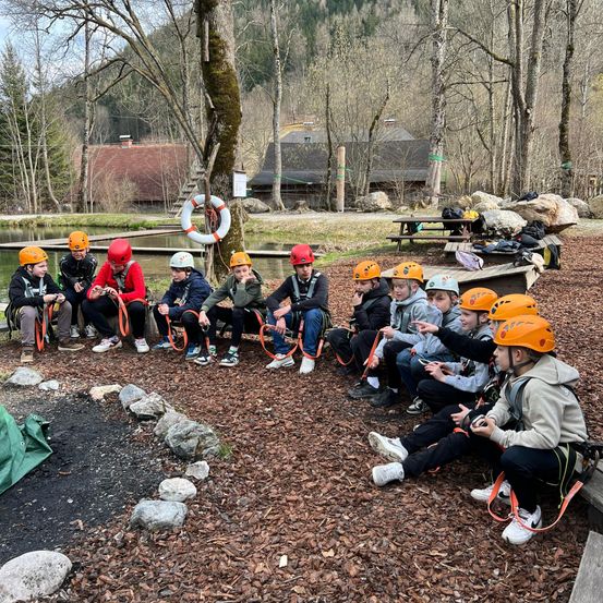 Eine Gruppe von Kindern mit orangefarbenen Helmen sitzt im Kreis in einem Waldgebiet, möglicherweise an einer Abenteueraktivität teilnehmend.