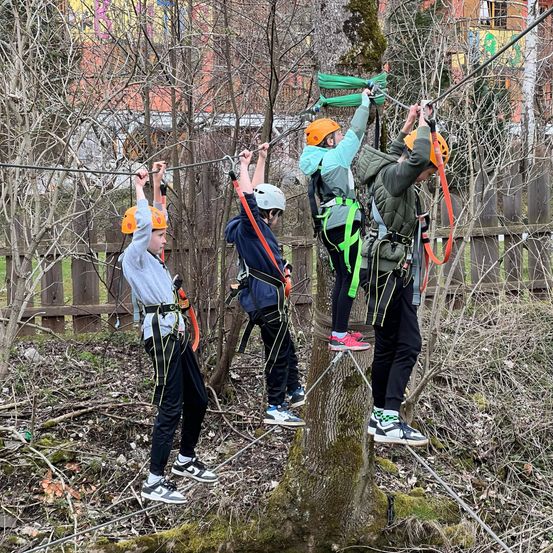 Vier Kinder gehen auf einem Seil, das an einem Baum im Wald gebunden ist. Sie tragen Sicherheitsgurte und Helme.