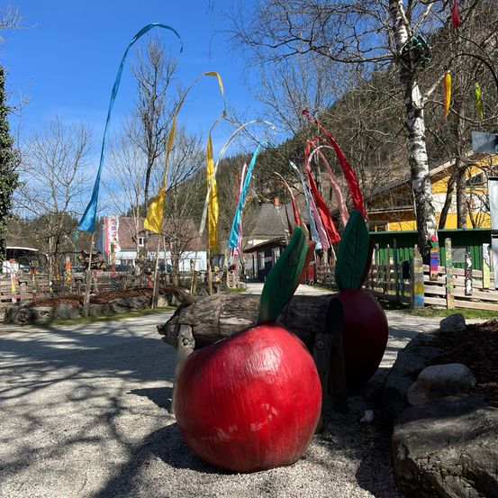Eine rote Apfelskulptur ruht auf Kiesboden unter einem klaren blauen Himmel, mit Bäumen, bunten Fahnen und einem gelben Gebäude in der Ferne mit einem Berg im Hintergrund.