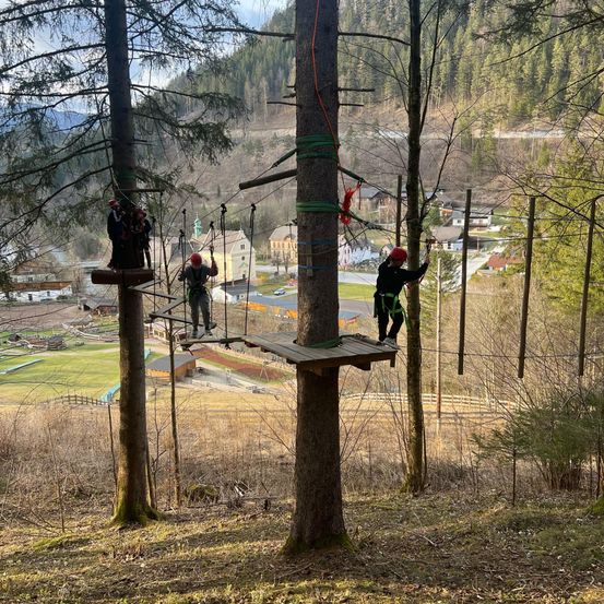 Vier Personen mit roten Helmen befinden sich auf einem Seilparcours in einem Wald, wobei sich zwei Personen auf Plattformen befinden und zwei andere an Seilen hängen. Ein Dorf mit Häusern und einer Kirche ist im Hintergrund zu sehen.