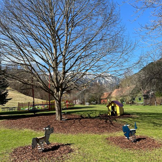 Ein Park mit einem Spielplatz, einem Baum und einem kleinen gelben Hundehäuschen. Ein Pferd und ein blauer Dinosaurier liegen auf dem Boden.