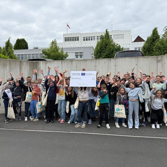 Eine Gruppe von Schülern posiert für ein Foto vor einem Gebäude mit einem Schild, das Interreg sagt. Sie halten einen großen Scheck und einige von ihnen halten Äpfel.