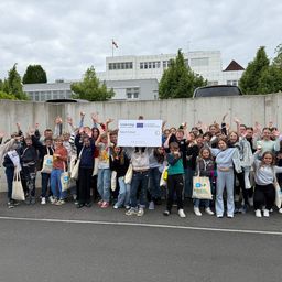 Eine Gruppe von Schülern posiert für ein Foto vor einem Gebäude mit einem Schild, das Interreg sagt. Sie halten einen großen Scheck und einige von ihnen halten Äpfel.