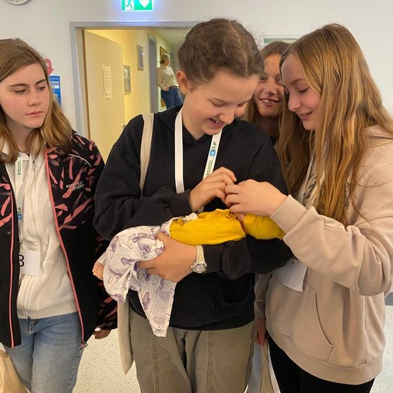 Vier junge Frauen sind drinnen, eine hält eine Babypuppe, während die anderen zuschauen, möglicherweise in einem Krankenhausflur.