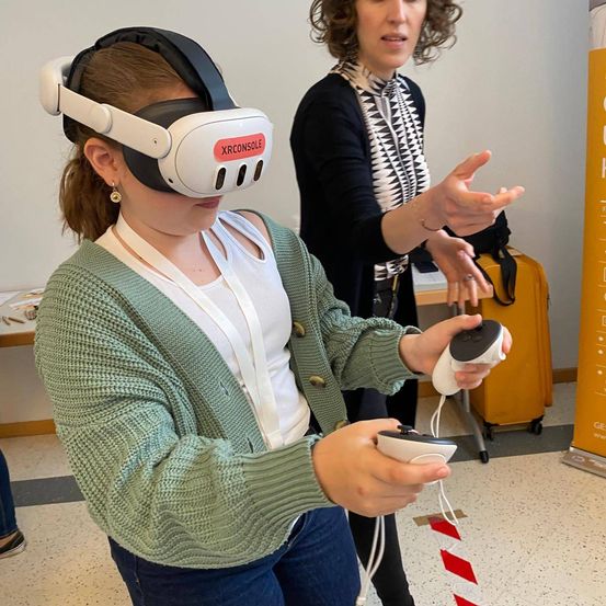 Eine Frau in einem grünen Strickjacke und Jeans trägt ein VR-Headset und hält einen Controller. Hinter ihr steht eine Frau mit lockigem Haar, die eine schwarz-weiße Jacke trägt und mit den Händen gestikuliert.