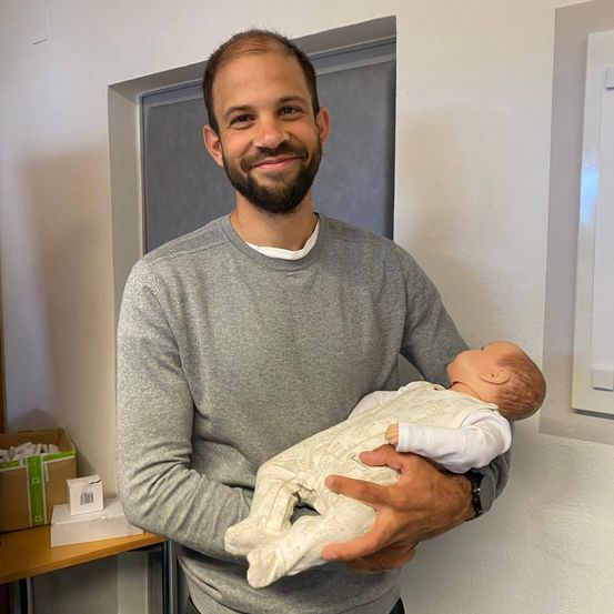 Ein Mann in einem grauen Langarmshirt hält eine Babypuppe mit lächelndem Gesicht in einem Raum.