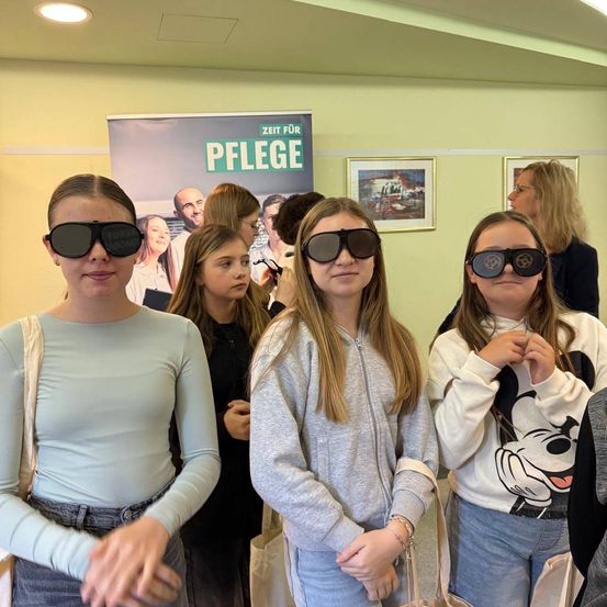 Vier Frauen stehen zusammen und tragen schwarze Sonnenbrillen. Dahinter ist ein Plakat mit dem Text Zeit fur Pflege. Eine weitere Frau steht im Hintergrund. Gerahmte Bilder hängen an der Wand.