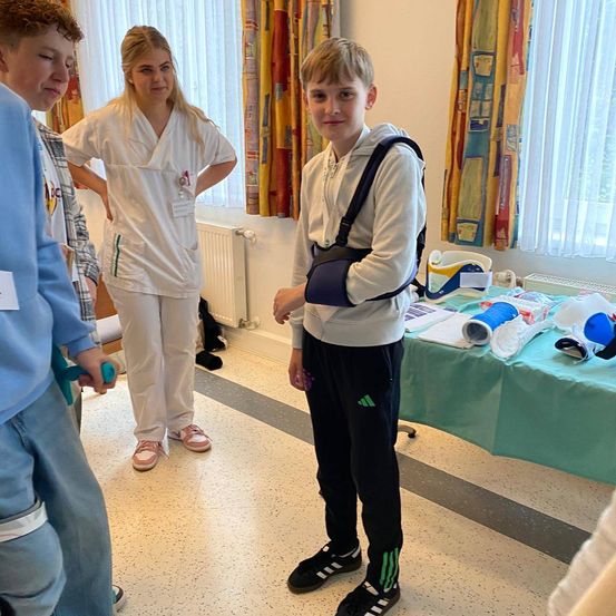 Ein junger Junge mit einem Arm in der Schlinge steht in einem Krankenhauszimmer mit einer Frau in Scrubs und einem Jungen mit einem grünen Stock.