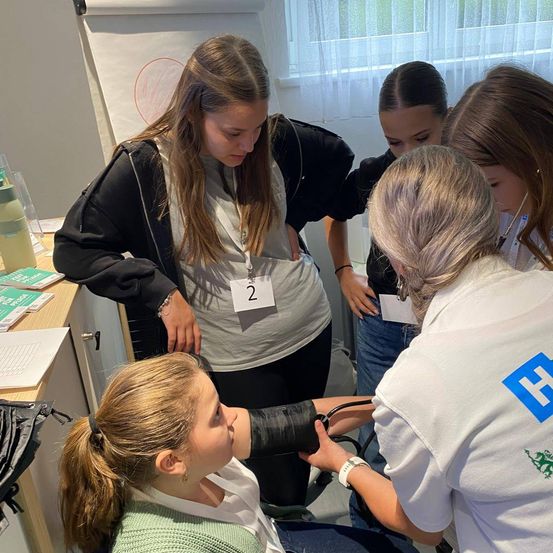 Fünf Frauen in einem Raum, eine misst den Blutdruck, zwei beobachten, eine trägt ein Namensschild. Bücher, ein Glas und eine Tafel mit einem roten Kreis im Hintergrund.