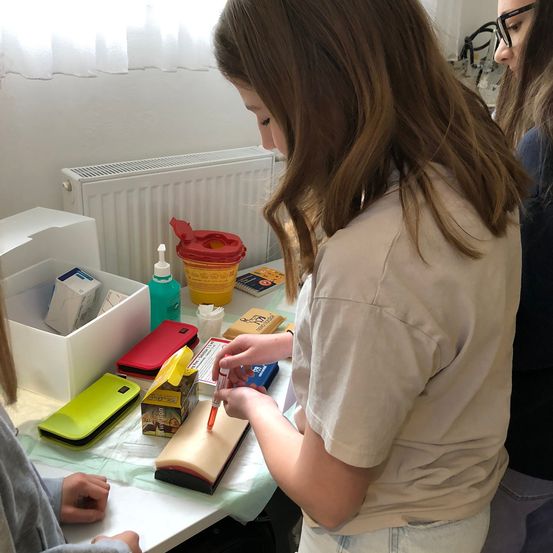 Ein Mädchen übt das Injizieren in einen künstlichen Arm mit einer Spritze, während andere zuschauen. Verschiedene medizinische Utensilien liegen auf dem Tisch.