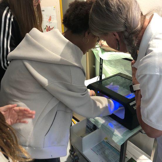 Eine Frau in einer weißen Kapuzenjacke und eine Frau in einem weißen Laborkittel untersuchen ein schwarzes elektronisches Gerät mit blauem Licht.