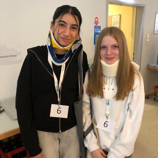 Zwei Mädchen mit Halskrausen mit der Nummer 6 stehen zusammen in einem Raum mit weißen Wänden. Eines trägt eine schwarze Jacke und das andere ein weißes Sweatshirt. Sie scheinen für ein Foto zu posieren.