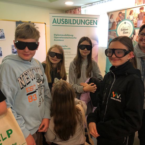 Eine Gruppe junger Personen, die Brillen tragen, lächelt und posiert für ein Foto vor einem Schild, das Ausbildungen anzeigt.