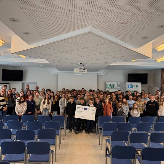 Eine große Gruppe von Schülern versammelt sich in einem Raum, einige halten einen großen Scheck. Sie stehen in Reihen von Stühlen. An den Wänden befinden sich Monitore und Schilder.