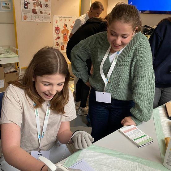 Zwei junge Frauen arbeiten an einem medizinischen Eingriff. Die Frau auf der linken Seite trägt Handschuhe und konzentriert sich auf die Aufgabe. Die Frau auf der rechten Seite lächelt und schaut auf die Aufgabe.
