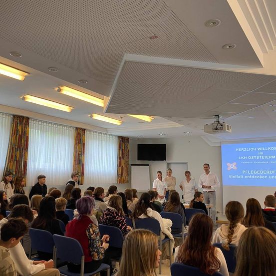 Ein Klassenzimmer voller Schüler, die in blauen Stühlen sitzen und einem Bildschirm gegenüberstehen. Mehrere Personen stehen vor dem Bildschirm, darunter zwei Männer und mehrere Frauen.