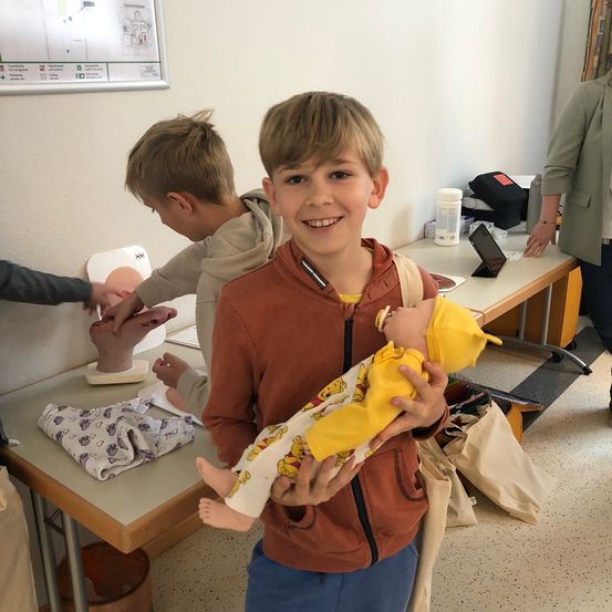 Ein junger Junge lächelt, während er eine Babypuppe in einem Klassenzimmer hält. Ein anderer Junge im Hintergrund interagiert mit einer Puppe.