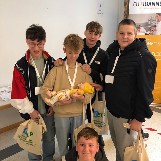 Fünf junge Jungen stehen zusammen und lächeln für ein Foto. Der Junge in der Mitte hält eine Babypuppe. Die Jungen tragen Freizeitkleidung, und einer trägt eine Brille. Sie tragen Taschen, und eine Werbung ist im Hintergrund sichtbar.