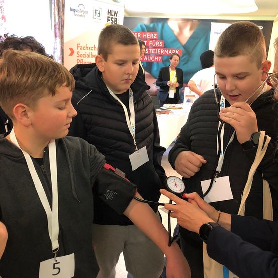 Drei junge Jungen sind an einem Gesundheitscheck-Stand. Ein Junge überprüft den Blutdruck eines anderen Jungen.
