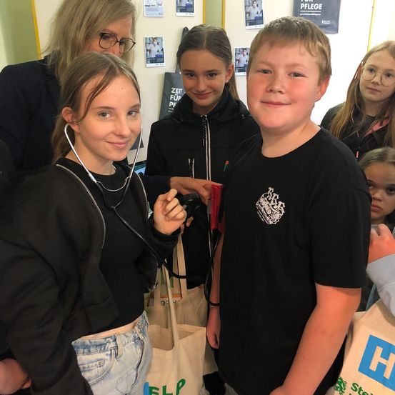 Eine Gruppe junger Menschen lächelt für ein Foto. Ein Mädchen hat ein Stethoskop um den Hals. Ein anderes Mädchen hält eine rote Mappe. Sie stehen eng beieinander.