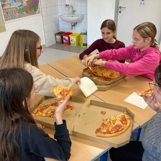 Fünf Mädchen sitzen an einem Tisch und essen Pizza. Ein Mädchen schneidet eine Pizza mit einem Messer.