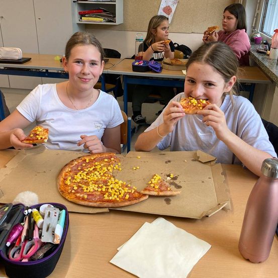 Zwei Mädchen sitzen an einem Tisch und essen Pizza. Eine von ihnen hält ein Stück Pizza mit Mais, während die andere ein Stück mit Käse isst.