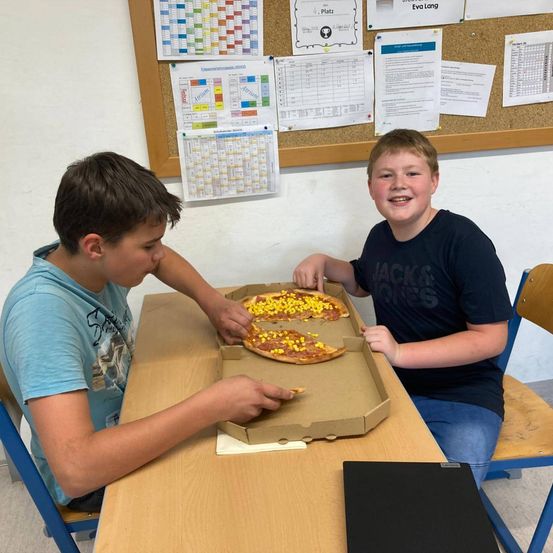 Zwei Jungen sitzen an einem Schreibtisch, einer holt sich eine Pizzascheibe und der andere lächelt. Auf dem Tisch steht eine Pizzaschachtel und ein Notizbuch.