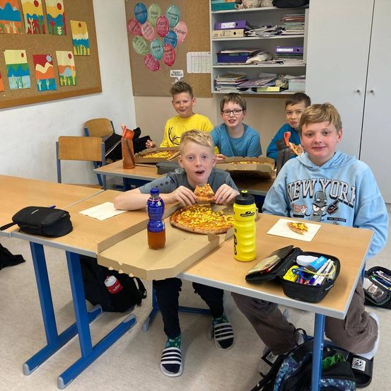 Fünf Jungen sitzen an einem Tisch im Klassenzimmer und essen Pizza aus Kartons. Zwei von ihnen tragen Hoodies, die anderen sind in Schuluniformen.