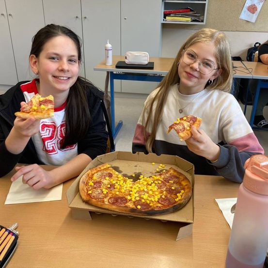 Zwei junge Mädchen sitzen in einem Klassenzimmer an einem Tisch und halten Stücke Pizza in den Händen. Auf dem Tisch steht eine Pizzaschachtel mit einer Pizza, die mit Mais und Peperoni belegt ist.