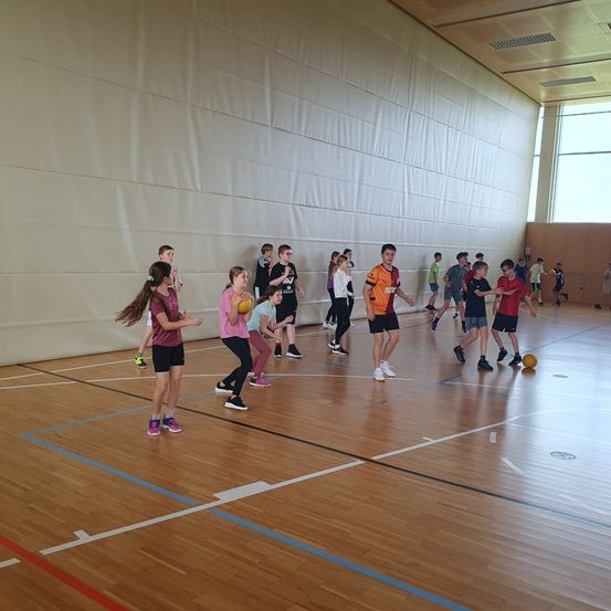Eine Gruppe von Kindern spielt Ball auf einem Hallenplatz. Einige von ihnen tragen Turnschuhe, andere sind barfuß.