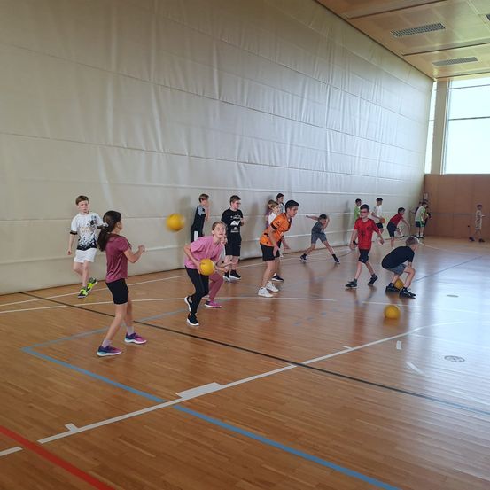 Eine Gruppe von Kindern spielt Dodgeball in einer Turnhalle. Sie halten alle Bälle und sind im Spiel engagiert.