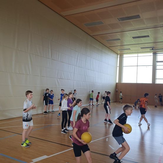 Eine Gruppe von Kindern und Jugendlichen spielt mit gelben Bällen auf einem Hallenspielfeld, einige stehen und andere sind in Bewegung. Das Spielfeld hat blaue Markierungen und eine weiße Wand mit einem weißen Vorhang.