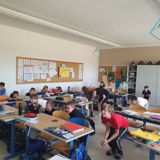 Ein Klassenzimmer mit mehreren Schülern, einige sitzen an Tischen, andere stehen, und einige lehnen sich an die Wand. Der Raum ist mit verschiedenen Gegenständen auf den Tischen und Pinnwänden mit angehefteten Papieren gefüllt.