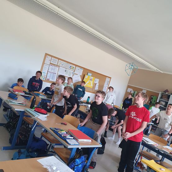 Eine Gruppe junger Jungen steht in einem Klassenzimmer. Die Schreibtische sind in Reihen angeordnet. Der Junge vorne trägt ein rotes T-Shirt und scheint zu sprechen.