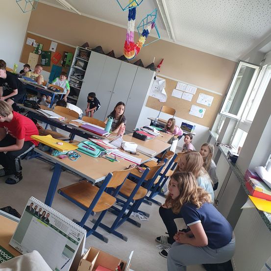 Ein Klassenzimmer mit Schülern, die an Tischen sitzen. Einige Schüler sitzen auf dem Boden und scheinen etwas zu hören. Es gibt Schränke, Poster und Dekorationen im Raum.