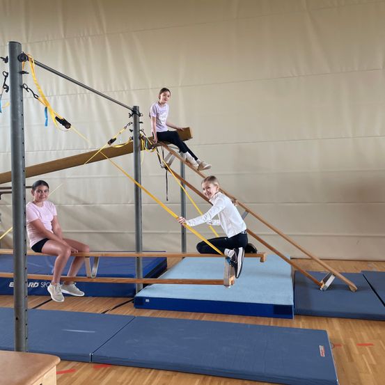 Drei Mädchen turnen an einer Barren in einer Turnhalle. Ein Mädchen springt, während die anderen beiden auf der Barren sitzen. Die Turnhalle hat Holzböden und blaue Matten.
