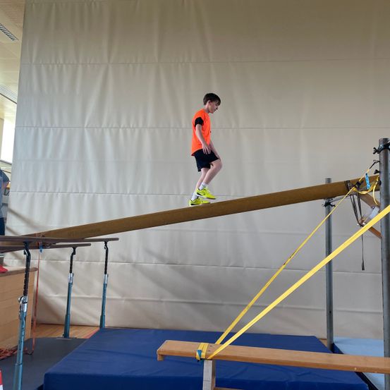 Ein junger Junge in einem orangefarbenen T-Shirt und schwarzen Shorts läuft auf einer Holzstange mit gelben Riemen. Er trägt gelbe Schuhe.