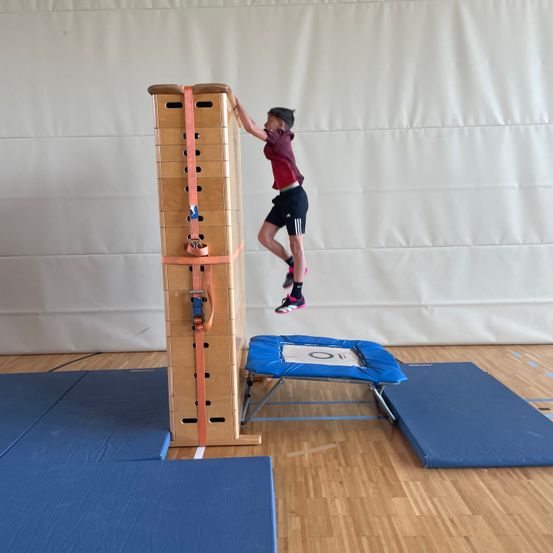 Ein junger Junge springt in einer Turnhalle auf einem Schaumstoffblock neben einer Holzkletterwand mit Gurten.