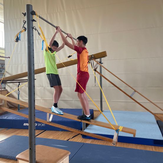Zwei Jungen in einem Fitnessstudio, die Klimmzüge an Parallelbarren durchführen. Ein Junge trägt ein grünes Shirt, der andere ein rotes.