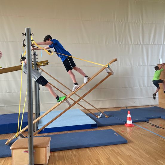 Drei junge Jungen nutzen ein Turngerät in einer Turnhalle mit blauen Matten, einem Kegel und einer großen weißen Wand.
