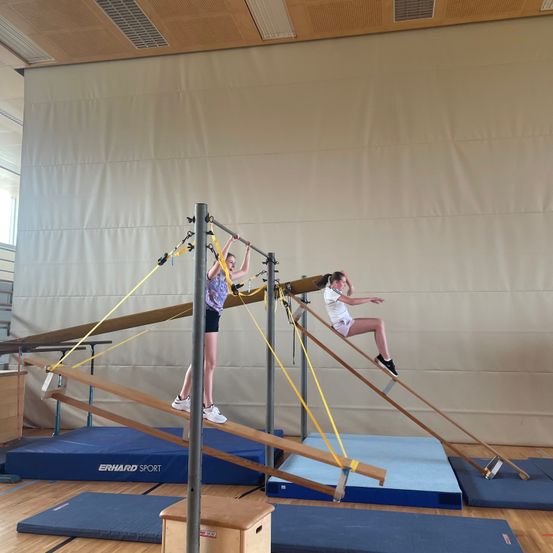 Zwei Mädchen trainieren am Reck, eine hält sich am Barren fest und die andere sitzt darauf. Der Raum hat einen Holzboden und eine große weiße Wand.