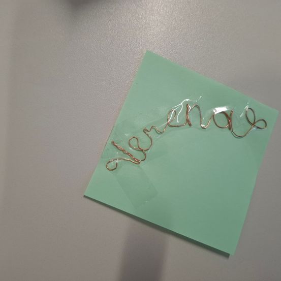 Ein grüner Haftnotizzettel mit Kupferdraht, der das Wort "Verena" bildet, befestigt durch einen transparenten Plastikstreifen.