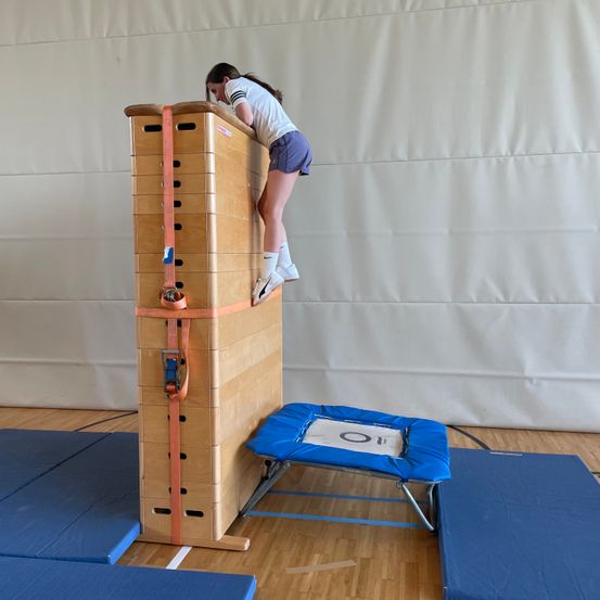 Ein junges Mädchen in einem weißen T-Shirt, Shorts und weißen Turnschuhen klettert eine hohe Holzkonstruktion mit einer blauen Matte an der Spitze.