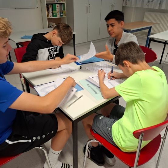 Vier junge Männer sitzen in einem Klassenzimmer um einen Tisch herum. Sie arbeiten an Papieren und einer hält einen Stift.
