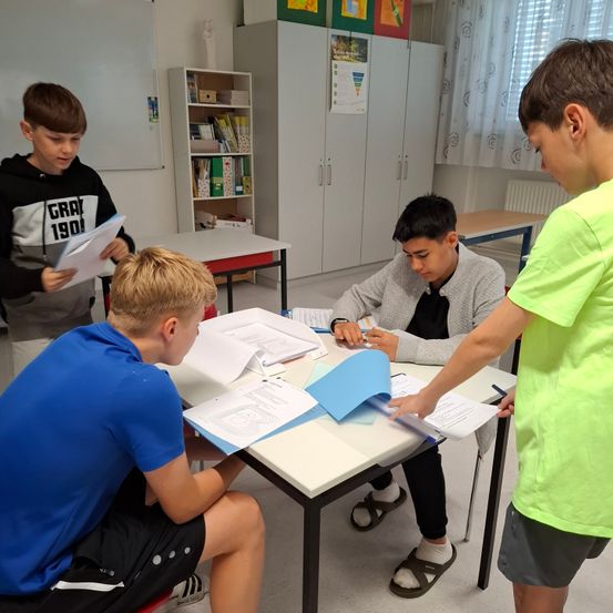Vier Jungen stehen in einem Klassenzimmer um einen Tisch herum. Sie betrachten ein Stück Papier auf dem Tisch, und einer von ihnen schreibt.