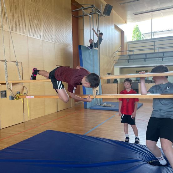 Drei Jungen führen Akrobatik in einem Fitnessstudio durch. Ein Junge hängt an einer Stange, ein anderer führt einen Handstand durch und der dritte beobachtet.