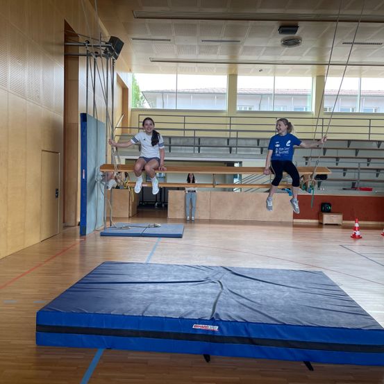 Zwei Mädchen führen Akrobatik auf einer horizontalen Stange in einer Turnhalle aus, mit einer blauen Matte auf dem Boden zur Sicherheit.