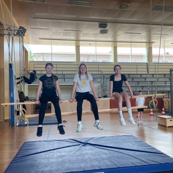 Drei junge Mädchen machen Gymnastik an einer horizontalen Stange in einer Turnhalle. Sie sitzen auf der Stange und lächeln.