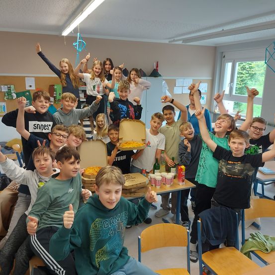 Eine Gruppe junger Schüler posiert für ein Foto im Klassenzimmer. Einige sitzen auf Tischen, andere stehen dahinter. Pizzaschachteln stehen auf dem Tisch, mit Bechern und Flaschen. Die Schüler heben ihre Hände und lächeln.