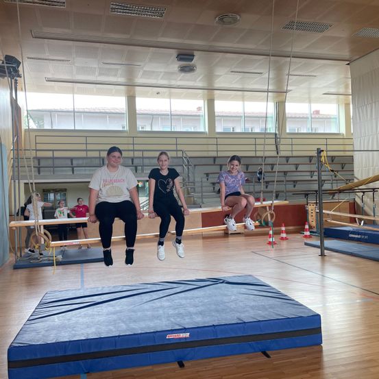 Drei Mädchen machen Akrobatik in einer Turnhalle. Eine hängt an einer Stange, und die anderen beiden machen einen Sprung. Die Turnhalle ist gut beleuchtet, und es gibt mehrere Übungsgeräte im Hintergrund.